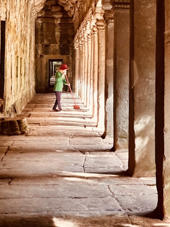 A person wearing a green shirt and red hat is sweeping the floor in a long, narrow corridor of an ancient stone structure. The corridor is lined with columns, and sunlight is streaming in, casting shadows. The stone floor and walls are weathered, adding a sense of antiquity.