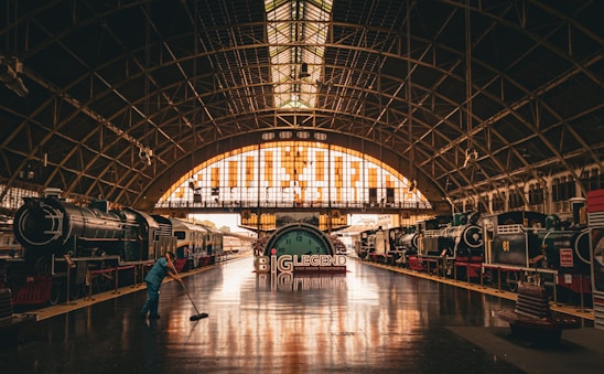 Inside a large, arched train station, vintage locomotives are displayed on both sides. A person in blue is cleaning the shiny, reflective floor. The station is illuminated by warm, orange-hued natural light streaming through the glass panels above.