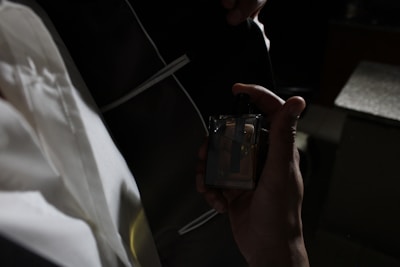 A person is holding a perfume bottle in one hand. The person is dressed in formal attire, including a white shirt and a black suit jacket with a white trim. The surroundings are dimly lit, creating a contrast between the illuminated areas and the dark background.