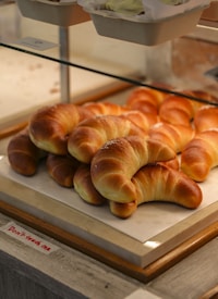 An assortment of golden-brown croissants arranged on a white tray inside a bakery display case. They have a slightly glossy finish, indicating freshness. Above the tray, other baked goods are visible in cardboard trays. A small sign below reads 'Don't touch me.'