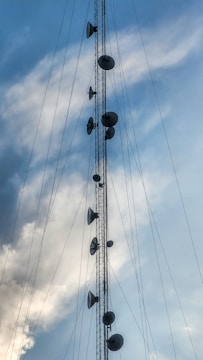 A tall communication tower with multiple satellite dishes and antennas is silhouetted against a cloudy sky. The structure is held in place by numerous cables, and the sky is predominantly blue with patches of white clouds.