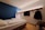 A modern hotel room features a single bed with white linens, adjacent to a blue accent wall. The space includes a wall-mounted flat-screen TV and a wooden rack with a hanger and coat. The ceiling lighting casts a warm, cozy ambiance, while a person's feet resting on the bed add a human touch.