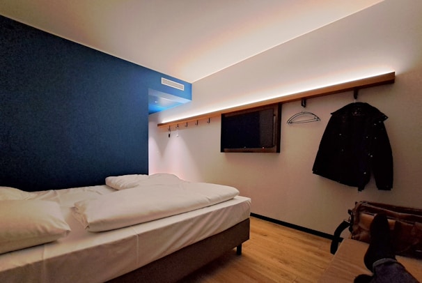 A modern hotel room features a single bed with white linens, adjacent to a blue accent wall. The space includes a wall-mounted flat-screen TV and a wooden rack with a hanger and coat. The ceiling lighting casts a warm, cozy ambiance, while a person's feet resting on the bed add a human touch.
