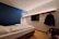 A modern hotel room features a single bed with white linens, adjacent to a blue accent wall. The space includes a wall-mounted flat-screen TV and a wooden rack with a hanger and coat. The ceiling lighting casts a warm, cozy ambiance, while a person's feet resting on the bed add a human touch.
