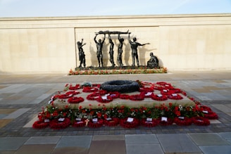 A solemn war memorial features a large stone slab covered with red poppy wreaths and surrounded by a paved area. Behind it, a bronze sculpture depicts several figures carrying a stretcher while another sits nearby, all against a backdrop of a wall inscribed with names.
