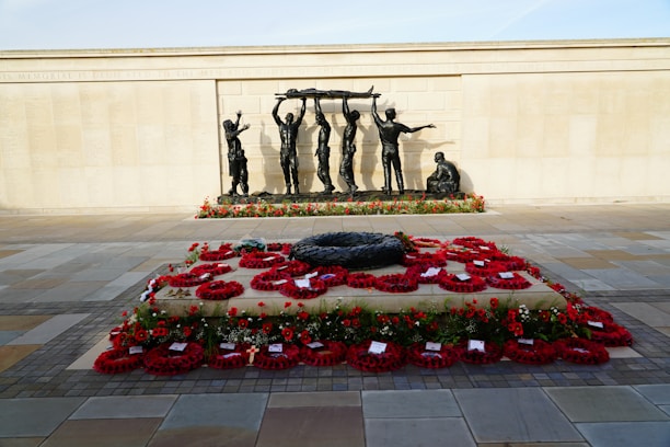 A solemn war memorial features a large stone slab covered with red poppy wreaths and surrounded by a paved area. Behind it, a bronze sculpture depicts several figures carrying a stretcher while another sits nearby, all against a backdrop of a wall inscribed with names.