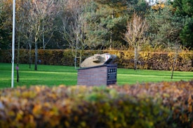 A model of a tank rests on a brick pedestal in a park setting. The tank is a small-scale replica, and the pedestal has an informational plaque on its side. Trees with bare branches and hedges with autumnal foliage fill the background, creating a serene and natural environment.