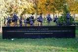 A bronze statue depicts a line of children carrying bags, symbolizing the evacuation of British children during World War II. The sculpture stands on a dark pedestal with an inscription dedicated to the memory of these events. The background features autumnal trees with vibrant yellow and green leaves.