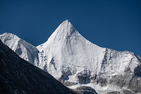 A majestic snow-covered mountain peak rises sharply under a clear blue sky. The texture of the snow and rock formations creates a striking contrast, highlighting the peak's rugged beauty.