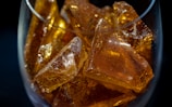 Amber-colored, translucent, crystalline-like pieces with smooth surfaces fill a glass container. The light reflecting off their surfaces creates a glowing effect.