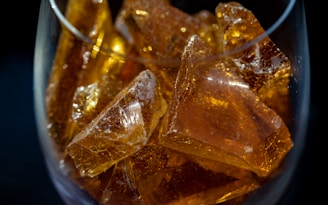 Amber-colored, translucent, crystalline-like pieces with smooth surfaces fill a glass container. The light reflecting off their surfaces creates a glowing effect.