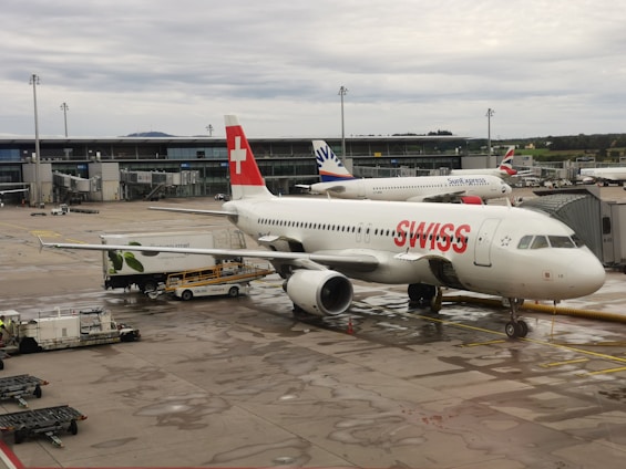 An airport scene featuring a SWISS airline aircraft stationed near a gate. The airplane is being serviced by ground crews with visible equipment around it, such as cargo loaders and fuel trucks. The tarmac is wet, indicating recent rainfall. In the background, other airplanes can be seen, including one from SunExpress. Modern airport infrastructure with terminal buildings is visible, and the sky is overcast.