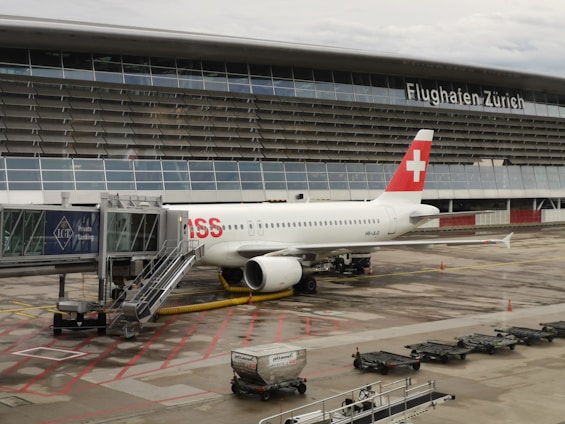 An airplane with a red and white tail featuring a cross symbol is parked at a gate at Zurich Airport. The aircraft is connected to a jet bridge, and there are several service vehicles and equipment nearby. The airport building, with its name displayed prominently, forms the backdrop.