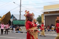 A group of people wearing ornate traditional costumes in red and gold, walking in a parade. Spectators sit on the sidewalk, watching the procession. The costumes include intricate headpieces adorned with flowers and lace. A large decorative wing is held by a participant, and various other people are dressed in casual clothing observing the event.