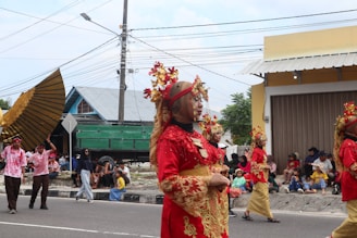 A group of people wearing ornate traditional costumes in red and gold, walking in a parade. Spectators sit on the sidewalk, watching the procession. The costumes include intricate headpieces adorned with flowers and lace. A large decorative wing is held by a participant, and various other people are dressed in casual clothing observing the event.