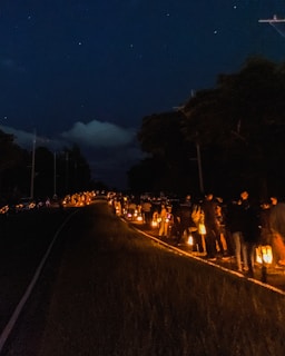 A group of people are walking along a dark road at night, each carrying a lantern that casts a warm glow. The scene is set under a starry sky, with trees lining the road.