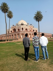 Three people stand on a grassy area facing a large, historic building with distinct Mughal architecture featuring a large dome and arches. Tall palm trees frame the scene, and the sky is clear with a few birds flying.
