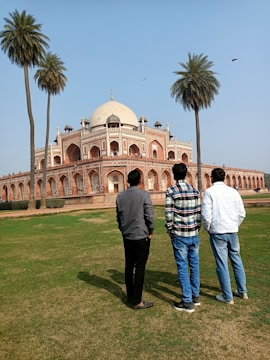 Three people stand on a grassy area facing a large, historic building with distinct Mughal architecture featuring a large dome and arches. Tall palm trees frame the scene, and the sky is clear with a few birds flying.