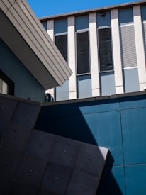 A modern architectural scene featuring intersecting geometric shapes and lines. The image highlights the contrast between vertical and diagonal elements, with a focus on concrete and metal surfaces. Tall windows are arranged in a symmetrical pattern, complemented by deep shadows and a clear blue sky.