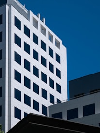 A modern white high-rise building with a grid-like pattern of dark windows against a clear blue sky. The building has clean lines and geometric shapes, suggesting an urban architectural style.