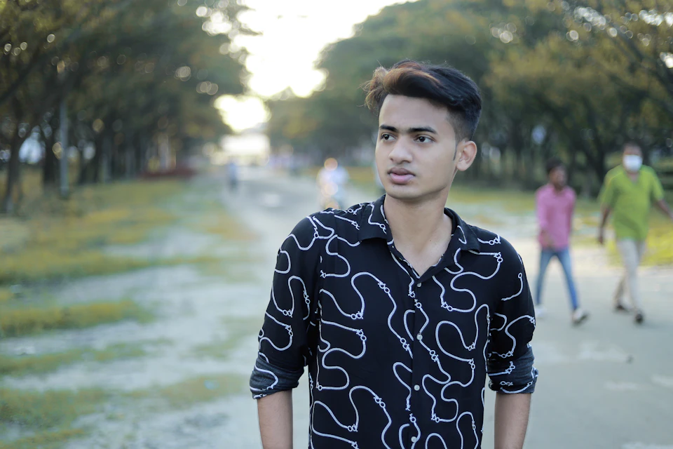 The image depicts a young man wearing a black and white shirt with white swirls, walking down a path surrounded by trees. The man is looking off to the side, possibly observing something or someone out of frame. In the background, there are other people present in the park setting, adding depth to the scene.

The image has a slightly blurred quality, giving it a dreamy and ethereal feel.