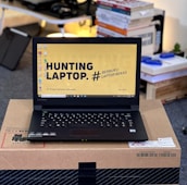 A laptop with an open screen displaying a desktop with a yellow background and bold text that says 'HUNTING LAPTOP.' It is placed on top of a cardboard box in a room with stacks of books and some electronic items in the background.