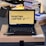 A laptop with an open screen displaying a desktop with a yellow background and bold text that says 'HUNTING LAPTOP.' It is placed on top of a cardboard box in a room with stacks of books and some electronic items in the background.