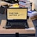 A laptop with an open screen displaying a desktop with a yellow background and bold text that says 'HUNTING LAPTOP.' It is placed on top of a cardboard box in a room with stacks of books and some electronic items in the background.