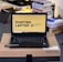 A laptop with an open screen displaying a desktop with a yellow background and bold text that says 'HUNTING LAPTOP.' It is placed on top of a cardboard box in a room with stacks of books and some electronic items in the background.