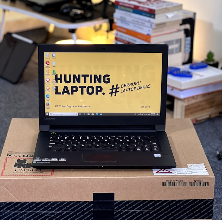 A laptop with an open screen displaying a desktop with a yellow background and bold text that says 'HUNTING LAPTOP.' It is placed on top of a cardboard box in a room with stacks of books and some electronic items in the background.