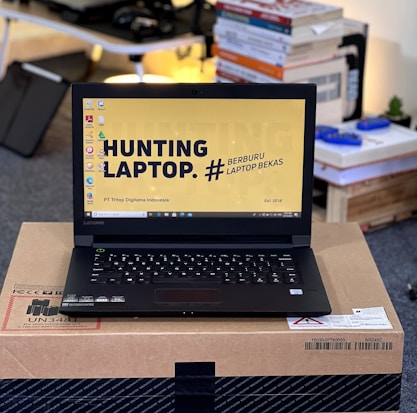 A laptop with an open screen displaying a desktop with a yellow background and bold text that says 'HUNTING LAPTOP.' It is placed on top of a cardboard box in a room with stacks of books and some electronic items in the background.