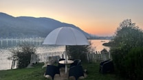 A serene lakeside setting with a table and black chairs under a large white umbrella, surrounded by greenery. The lake reflects the soft hues of the sunset, and mountains frame the background. A white fence partially encloses the area, giving it a cozy feel.