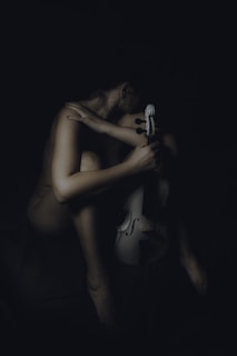 A person is sitting in a dark setting, holding a white violin. The posture suggests deep thought or introspection, with the person's arm wrapped around their shoulder and head turned slightly downwards.