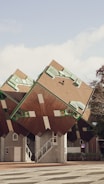A group of unique, cube-shaped houses tilted at an unusual angle, featuring geometric windows with green frames and brown slanted roofs. The setting includes a small staircase leading up to one of the structures and a nearby tree with some foliage visible. A bicycle is parked near the entrance area of one of the houses, adding a touch of urban life to the architectural scene.