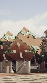 A group of unique, cube-shaped houses tilted at an unusual angle, featuring geometric windows with green frames and brown slanted roofs. The setting includes a small staircase leading up to one of the structures and a nearby tree with some foliage visible. A bicycle is parked near the entrance area of one of the houses, adding a touch of urban life to the architectural scene.
