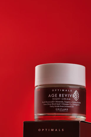 A jar of Age Revive night cream from Oriflame is placed against a bold red background. The jar is transparent with a white lid, featuring text detailing its benefits and ingredients. It rests on a dark box with brand titling.