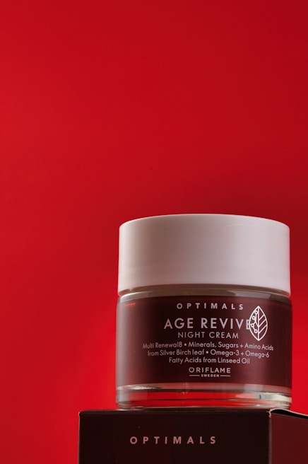 A jar of Age Revive night cream from Oriflame is placed against a bold red background. The jar is transparent with a white lid, featuring text detailing its benefits and ingredients. It rests on a dark box with brand titling.