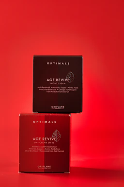 Two skincare product boxes labeled as Age Revive by Oriflame are stacked against a vibrant red background. The upper box is a darker shade of red, labeled for night cream, while the lower box is a lighter shade, labeled for day cream with SPF 15.