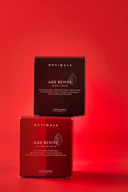 Two skincare product boxes labeled as Age Revive by Oriflame are stacked against a vibrant red background. The upper box is a darker shade of red, labeled for night cream, while the lower box is a lighter shade, labeled for day cream with SPF 15.