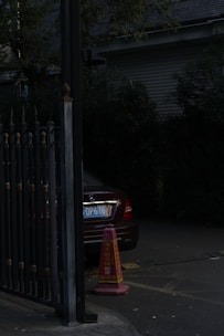 A partially visible car with a visible license plate is parked next to a metal gate. A traffic cone with writing on it is in front of the car. The scene is dimly lit, and there are trees and a building in the background.