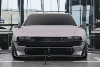 A sleek, futuristic car with a matte gray finish is showcased in a modern, glass-walled showroom. The vehicle features angular lines, a low chassis, and wide tires, emphasizing a performance-oriented design.