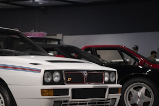 A close-up view of two vintage sports cars parked side by side indoors. The car in the foreground is white with blue and red racing stripes on the hood, featuring round headlights and a distinctive square grille. Next to it is a car with a red roof and black body. The setting appears to be a showroom or museum with subdued lighting and a dark ceiling.