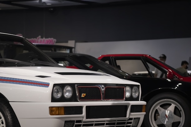 A close-up view of two vintage sports cars parked side by side indoors. The car in the foreground is white with blue and red racing stripes on the hood, featuring round headlights and a distinctive square grille. Next to it is a car with a red roof and black body. The setting appears to be a showroom or museum with subdued lighting and a dark ceiling.