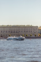 A hydrofoil boat is moving on a body of water in front of a large, classical-style building. The building is adorned with numerous windows and a green roof, and it is lined with trees on the waterfront. The clear sky in the background suggests a pleasant day.