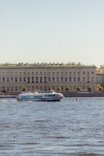 A hydrofoil boat is moving on a body of water in front of a large, classical-style building. The building is adorned with numerous windows and a green roof, and it is lined with trees on the waterfront. The clear sky in the background suggests a pleasant day.