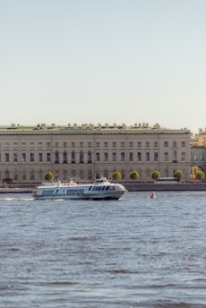 A hydrofoil boat is moving on a body of water in front of a large, classical-style building. The building is adorned with numerous windows and a green roof, and it is lined with trees on the waterfront. The clear sky in the background suggests a pleasant day.