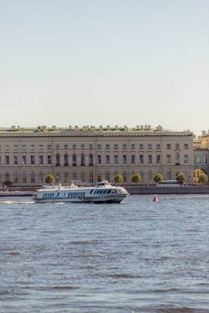 A hydrofoil boat is moving on a body of water in front of a large, classical-style building. The building is adorned with numerous windows and a green roof, and it is lined with trees on the waterfront. The clear sky in the background suggests a pleasant day.