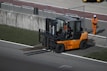 A forklift with an orange body and black details is positioned near a concrete barrier with two numbered signs. The operator inside the forklift is wearing a helmet. Nearby, a person in an orange jumpsuit and white safety helmet is standing on a paved surface. The area is bordered by grass and contains metallic railings.