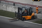 A forklift with an orange body and black details is positioned near a concrete barrier with two numbered signs. The operator inside the forklift is wearing a helmet. Nearby, a person in an orange jumpsuit and white safety helmet is standing on a paved surface. The area is bordered by grass and contains metallic railings.