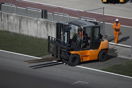 A forklift with an orange body and black details is positioned near a concrete barrier with two numbered signs. The operator inside the forklift is wearing a helmet. Nearby, a person in an orange jumpsuit and white safety helmet is standing on a paved surface. The area is bordered by grass and contains metallic railings.